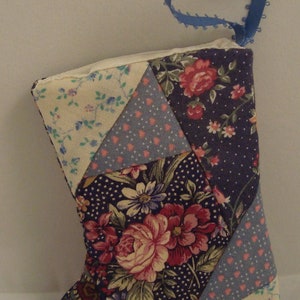 May include: A patchwork fabric stocking with a blue ribbon loop at the top. The stocking is made of various floral patterns in shades of pink, blue, and white on a dark purple background.