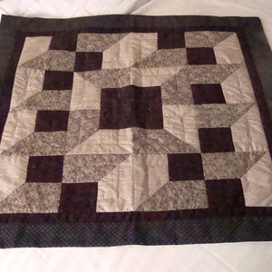 May include: A square quilt with a geometric pattern in shades of brown and beige. The quilt is made of fabric squares sewn together to create a repeating pattern. The quilt is edged with a dark brown border.