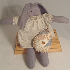 May include: A purple bunny doll wearing a white dress with a floral pattern and holding a patchwork egg ornament. The bunny is sitting on a wooden crate.