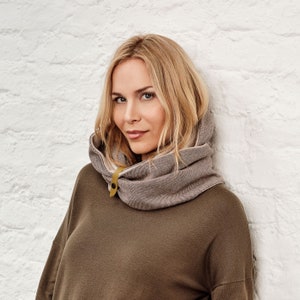 May include: A gray and beige cowl neck scarf with a yellow button closure. The scarf is made of a soft, textured fabric and is worn over a dark green long-sleeve shirt. The background is a white brick wall.