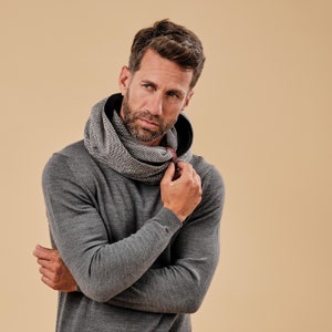 Puede incluir: Un hombre con un suéter gris y una bufanda de patrón blanco y negro. La bufanda tiene una correa de cuero marrón.