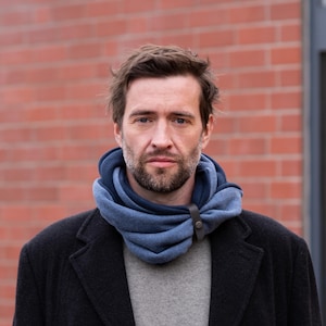 Può includere: Una persona indossa una sciarpa con cappuccio blu e grigia con chiusura in pelle. La sciarpa è a strati e sembra essere fatta di un materiale morbido. La persona indossa anche un maglione grigio e un cappotto scuro.