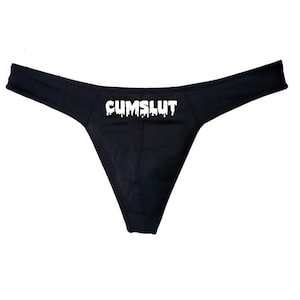 May include: A black thong featuring the text CUMSLUT printed in a white, dripping, liquid-style font across the front centre. The garment has a thin waistband and a triangular front panel, maintaining a solid black colour throughout the design.