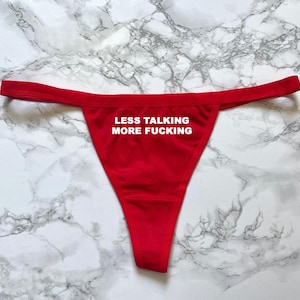 Könnte beinhalten: Roter String-Tanga mit dem weißen Text "LESS TALKING MORE FUCKING". Die Unterwäsche wird auf einer marmorierten Oberfläche präsentiert.