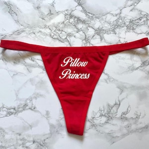 May include: Red thong underwear with the text "Pillow Princess" in a white script. The underwear is displayed on a marble surface. The thong is a solid red color.