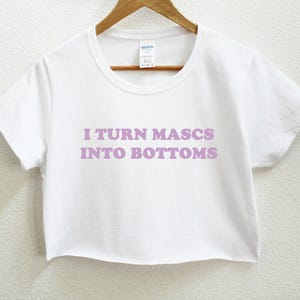 May include: White crop top t-shirt with the text "I TURN MASCS INTO BOTTOMS" in lavender lettering. The shirt is hanging on a wooden hanger against a white background. The shirt has a crew neck and short sleeves.