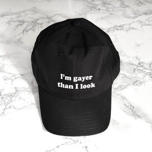 Peut inclure: Une casquette de baseball noire avec du texte blanc qui dit "Je suis plus gay que je n'en ai l'air".
