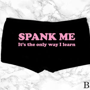 May include: Black boxer shorts with the text "SPANK ME" in pink, followed by "It's the only way I learn". The word "Back" is printed at the bottom. The shorts are displayed on a white and grey marble surface.