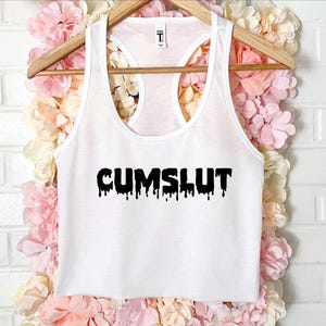 May include: White racerback vest with the word "CUMSLUT" printed in black, dripping font. The vest is hanging on a wooden hanger against a backdrop of pink and white flowers.
