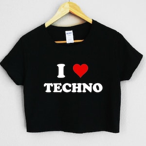 Puede incluir: Top corto negro con el texto "I ❤️ TECHNO" en blanco, con un símbolo de corazón rojo. La camiseta está colgada de una percha de madera sobre un fondo blanco. La camiseta está hecha de un material suave.