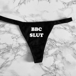 May include: Black thong underwear with the text "BBC SLUT" in white block letters. The underwear is displayed on a white and gray marble patterned surface.