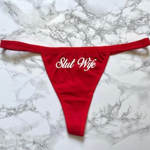 May include: A bright red thong featuring the text Slut Wife printed in a white cursive script across the front centre. The garment has a thin waistband and a triangular front panel, constructed from a smooth, solid-coloured fabric material.