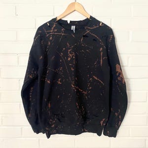 May include: Black sweatshirt with brown bleach splatters and distressed holes. The sweatshirt has long sleeves and a crew neck.
