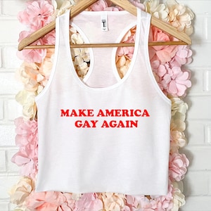 Könnte beinhalten: Weißes Tanktop mit dem roten Text "MAKE AMERICA GAY AGAIN". Das Oberteil hängt an einem Holzbügel vor einem Hintergrund aus rosa und weißen Blumen. Das Tanktop hat ein Racerback-Design.