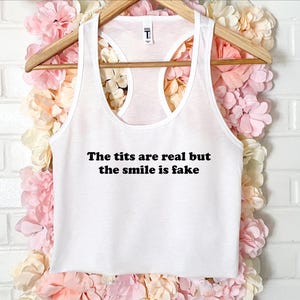 May include: White racerback tank top with the text "The tits are real but the smile is fake" in black. The tank top is hanging on a wooden hanger against a backdrop of pink and cream-colored flowers.