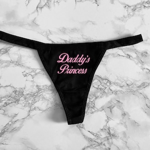 May include: A black thong featuring the text Daddy's Princess printed in a pink script font across the front center. The garment has a thin waistband and a triangular front panel, constructed from a smooth, solid black fabric material.
