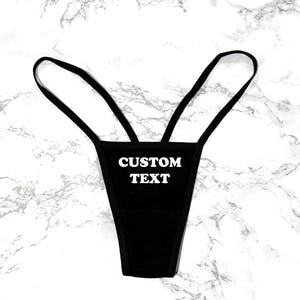 May include: A black thong featuring thin side straps and the words CUSTOM TEXT printed in white capital letters on the front center. The garment has a minimalist design with a solid black fabric construction and a high-cut leg opening.