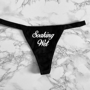May include: A black thong featuring the white text Soaking Wet printed in a cursive script font on the front center. The garment has a thin waistband and a triangular front panel, constructed from a solid black fabric with a smooth finish.
