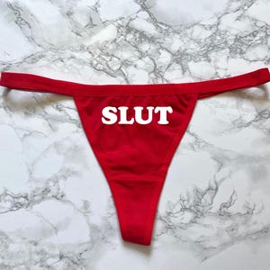 May include: Red thong underwear with the word "SLUT" printed in white across the front. The underwear is displayed on a marble-patterned surface.