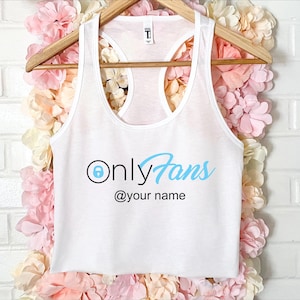May include: White racerback vest with the text "OnlyFans" in light blue and black, with a padlock icon. The text "@your name" is below the main text. The vest is hanging on a wooden hanger.
