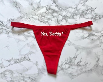 Puede incluir: Tanga rojo con el texto "Yes, Daddy?" en escritura blanca. La ropa interior se muestra sobre una superficie con vetas de mármol.