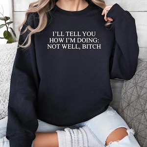 Peut inclure: Sweat-shirt noir avec l'inscription blanche "I'LL TELL YOU HOW I'M DOING: NOT WELL, BITCH". Le sweat-shirt est de style col rond. La personne porte un jean bleu clair.