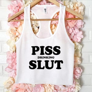 Può includere: Canotta bianca con le parole "PISS DRINKING SLUT" stampate in grassetto nero. La canotta è appesa a una gruccia di legno su uno sfondo di fiori rosa e bianchi.