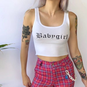 May include: White crop top with a black "Babygirl" graphic in a gothic font. The top is worn with a pink and white plaid skirt and a silver chain with a blue dice charm.