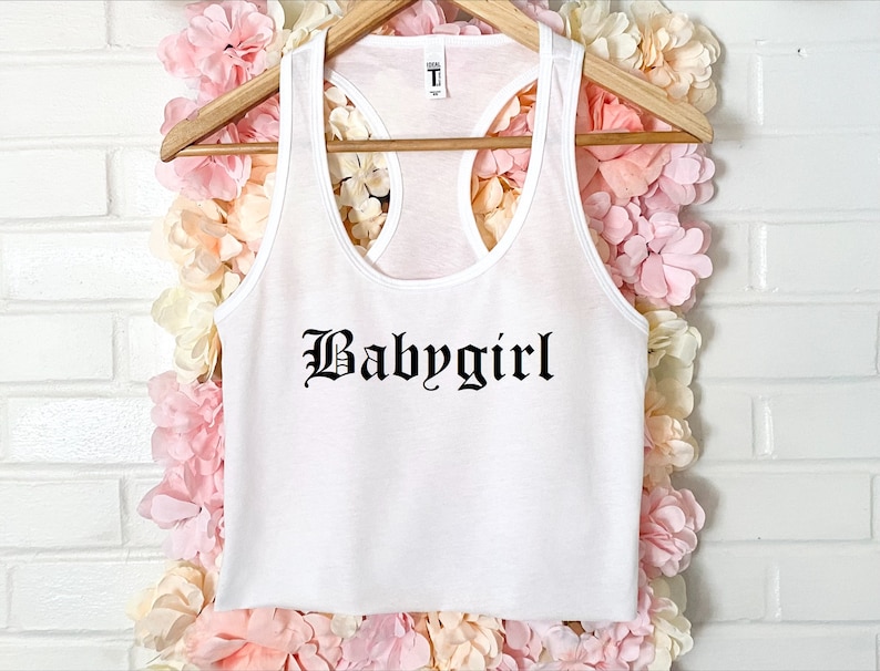 May include: White tank top with a black "Babygirl" graphic in a gothic font.