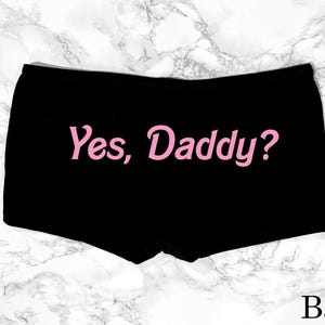 Op de afbeelding: Zwarte boxershort met de tekst "Yes, Daddy?" in roze letters. Het kledingstuk wordt gepresenteerd tegen een achtergrond met marmerpatroon. Het woord "Back" is onderaan zichtbaar.