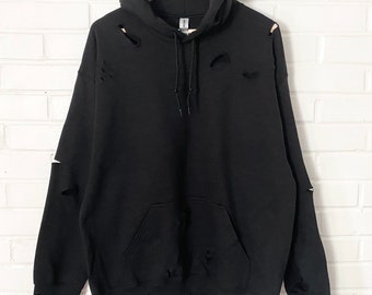 May include: A black long-sleeve hooded sweatshirt featuring a distressed design with multiple small rips and tears across the chest, shoulders, sleeves, and hem. The garment includes a front kangaroo pocket, a drawstring hood, and ribbed cuffs.