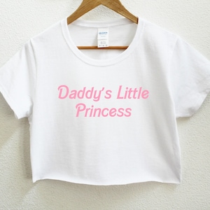 May include: White crop top with the pink text "Daddy's Little Princess". The t-shirt is hanging on a wooden hanger. The shirt is made of a soft material.
