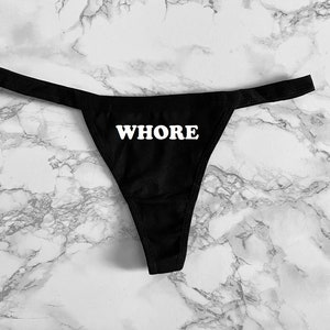 Pode incluir: Tanga de renda preta com a palavra "WHORE" impressa em branco na frente.