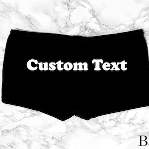 Pode incluir: Boxer preto com o texto "Custom Text" em letras brancas na frente. A palavra "Back" é impressa em branco na parte inferior. O fundo é um padrão de mármore.