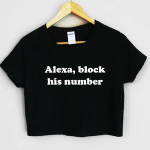 May include: Black crop top with the text "Alexa, block his number" in white. The shirt is hanging on a wooden hanger against a white background. The shirt is a casual style.