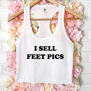 May include: White racerback tank top with the text "I SELL FEET PICS" in bold black letters. The tank top is hanging on a wooden hanger against a backdrop of pink and cream-colored flowers.