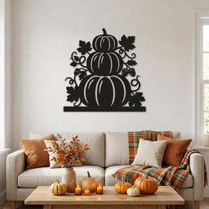 May include: A black metal wall art piece featuring a stack of three pumpkins surrounded by decorative leaves and acorns. The design is flat, silhouette-style, and mounted on a wall above a sofa, showcasing a clean, symmetrical arrangement.