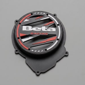 Peut inclure: Un couvercle d'embrayage moteur circulaire noir avec un design ventilé et des accents rouges et blancs. Le centre affiche le texte blanc Beta et le texte rouge RACING, avec le mot VOCA imprimé en haut et en bas du rebord circulaire extérieur.