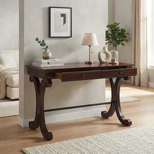 May include: A dark brown wooden writing desk featuring two drawers with small round knobs, supported by curved legs connected by a horizontal metal bar. The desk surface holds a stack of books, a small vase, a table lamp, and a ceramic sculpture.