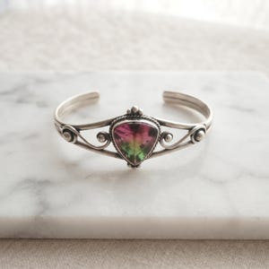 Peut inclure: Un bracelet manchette en argent orné d'une pierre précieuse centrale en tourmaline melon d'eau triangulaire aux teintes roses et vertes. La bande métallique présente des détails en fil torsadé autour de la pierre et des volutes argentées.