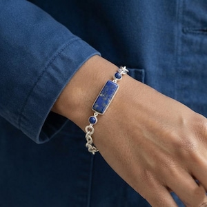 May include: A silver chain bracelet featuring a central rectangular lapis lazuli stone and two smaller round lapis lazuli stones on either side. The bracelet is constructed with polished silver links and a secure clasp mechanism visible on the wrist.