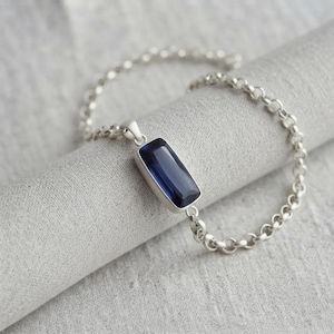 May include: A silver chain bracelet featuring a single rectangular, faceted deep blue gemstone pendant. The pendant is set in a smooth silver frame, and the bracelet is constructed from polished round silver links that form a continuous chain.