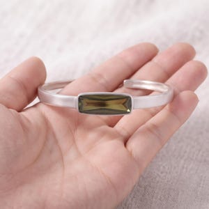 May include: A matte silver-toned cuff bracelet featuring a central rectangular, faceted olive-green gemstone. The bracelet has a smooth, open-ended band design with a polished finish on the inner surface and a muted, brushed texture on the exterior.