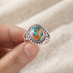 Può includere: Un anello in argento con una pietra ovale caratterizzata da un motivo a mosaico di segmenti blu brillante e arancioni. La pietra è circondata da un bordo perlato e da un dettaglio a corda intrecciata, su una fascia con volute traforate.