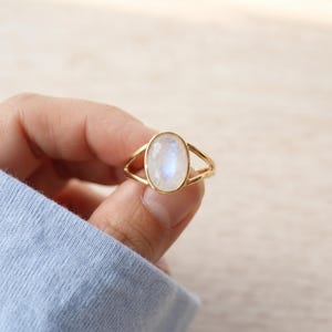 May include: A gold ring featuring an oval-shaped moonstone cabochon set in a polished metal band. The stone displays a soft, iridescent blue glow against the warm yellow tone of the gold setting, which includes a split shank design.