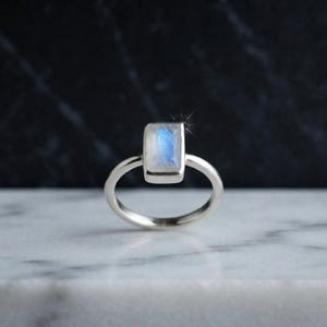 May include: A silver ring featuring a rectangular moonstone set in a simple metal band. The stone displays a translucent, milky white appearance with a distinct blue adularescence or flash of light emanating from the center of the gemstone.