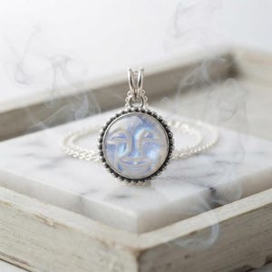 May include: A silver pendant necklace featuring a round, translucent moonstone carved with a smiling face. The stone is set in a beaded silver frame with a decorative bail, attached to a delicate silver chain, surrounded by wisps of white smoke.