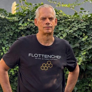 Könnte beinhalten: Ein schwarzes T-Shirt mit kurzen Ärmeln, das den weißen Schriftzug FLOTTENCHEF, den Text 44.000 MITARBEITERINNEN und eine gelbe Grafik einer Wabenstruktur aus sechs verbundenen Sechsecken im Brustbereich des Kleidungsstücks aufweist.