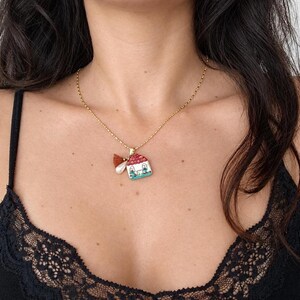Può includere: Una collana a catena color oro con un piccolo ciondolo a forma di casa con tetto rosso, pareti bianche e dettagli blu. Il ciondolo è accompagnato da una piccola perla bianca e da una minuscola nappa marrone appese allo stesso anello dorato.