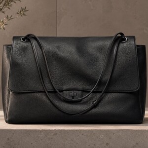 Könnte beinhalten: Eine schwarze Handtasche mit strukturierter Oberfläche, ausgestattet mit einer großen Frontklappe, einem rechteckigen Drehverschluss und zwei dünnen Schulterriemen. Die Tasche hat eine strukturierte rechteckige Form mit abgerundeten Ecken.